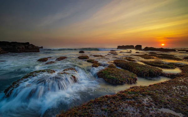 İnanılmaz bir deniz manzarası. Hareket eden dalgalı okyanus. Düşük lide. Yeşil yosun ve yosunlarla kaplı taşlar. Güneş ufukta görünüyor. Günbatımı manzara arka planı. Uzun pozlama. Yumuşak odaklanma. Mengening Plajı, Bali, Endonezya