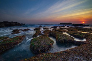 İnanılmaz bir deniz manzarası. Hareket eden dalgalı okyanus. Düşük lide. Yeşil yosun ve yosunlarla kaplı taşlar. Güneş ufukta görünüyor. Günbatımı manzara arka planı. Uzun pozlama. Yumuşak odaklanma. Mengening Plajı, Bali, Endonezya