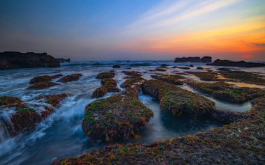 İnanılmaz bir deniz manzarası. Hareket eden dalgalı okyanus. Düşük lide. Yeşil yosun ve yosunlarla kaplı taşlar. Doğa geçmişi kavramı. Gün batımı manzarası. Uzun pozlama. Yumuşak odaklanma. Mengening Plajı, Bali, Endonezya