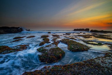 İnanılmaz bir deniz manzarası. Hareket eden dalgalı okyanus. Düşük lide. Yeşil yosun ve yosunlarla kaplı taşlar. Doğa geçmişi kavramı. Gün batımı manzarası. Uzun pozlama. Yumuşak odaklanma. Mengening Plajı, Bali, Endonezya