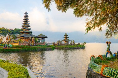 Bali adasındaki Pura Ulun Danu Bratan tapınağı. Güzel bale tapınağı. Bali 'nin simgesi. Bulutlu gökyüzü. Su yansıması. Ağaç dalları olan bir ön plan. Bratan Gölü, Bedugul, Bali, Endonezya