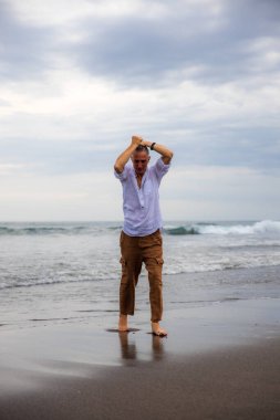 Sahilde yalınayak duran yalnız bir adam. Kahverengi pantolon ve beyaz gömlek giyen bir adam. Horizon deniz manzarası. Bulutlu gökyüzü. Seyahat konsepti. Uzayı kopyala Asya 'da tatil. Bali, Endonezya