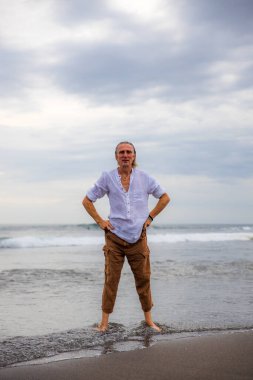 Sahilde yalınayak duran yalnız bir adam. Kahverengi pantolon ve beyaz gömlek giyen bir adam. Horizon deniz manzarası. Bulutlu gökyüzü. Seyahat konsepti. Uzayı kopyala Asya 'da tatil. Bali, Endonezya