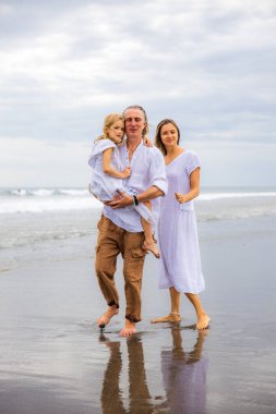 Mutlu aile kumsalda vakit geçiriyor. Yaz tatili. Aile tatili. Anne, baba ve kız sahilde yalınayak yürüyor. Baba, kızını kollarında taşıyor. Uzayı kopyala Bali, Endonezya