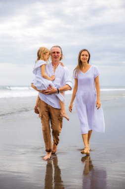Mutlu aile kumsalda vakit geçiriyor. Yaz tatili. Aile tatili. Anne, baba ve kız sahilde yalınayak yürüyor. Baba, kızını kollarında taşıyor. Uzayı kopyala Bali, Endonezya