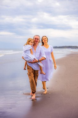 Mutlu aile kumsalda vakit geçiriyor. Yaz tatili. Aile tatili. Anne, baba ve kız sahilde yalınayak yürüyor. Baba, kızını kollarında taşıyor. Uzayı kopyala Bali, Endonezya