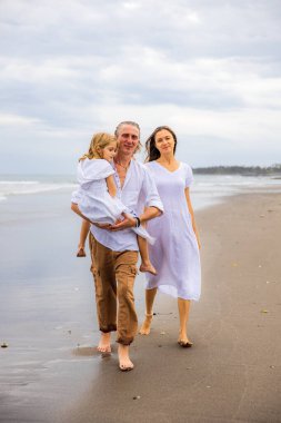 Mutlu aile kumsalda vakit geçiriyor. Yaz tatili. Aile tatili. Anne, baba ve kız sahilde yalınayak yürüyor. Baba, kızını kollarında taşıyor. Uzayı kopyala Bali, Endonezya