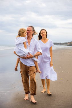 Mutlu aile kumsalda vakit geçiriyor. Yaz tatili. Aile tatili. Anne, baba ve kız sahilde yalınayak yürüyor. Baba, kızını kollarında taşıyor. Uzayı kopyala Bali, Endonezya