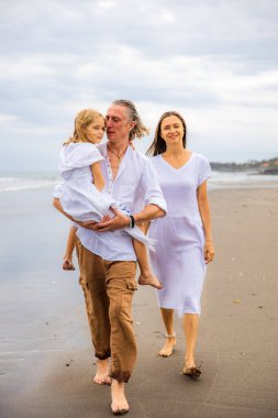 Mutlu aile kumsalda vakit geçiriyor. Yaz tatili. Aile tatili. Anne, baba ve kız sahilde yalınayak yürüyor. Baba, kızını kollarında taşıyor. Uzayı kopyala Bali, Endonezya