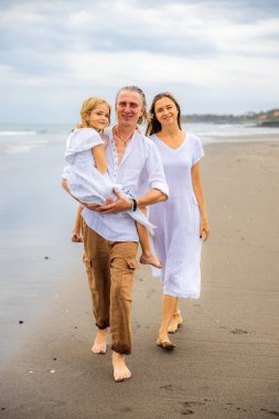 Mutlu aile kavramı. Yaz tatili. Asya 'da aile tatili. Anne, baba ve kızı sahilde yalınayak yürüyor. Baba, kızını kollarında taşıyor. Uzayı kopyala Bali, Endonezya