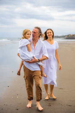 Mutlu aile kavramı. Yaz tatili. Asya 'da aile tatili. Anne, baba ve kızı sahilde yalınayak yürüyor. Baba, kızını kollarında taşıyor. Uzayı kopyala Bali, Endonezya