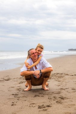 Kızı babaya sarılıyor. Kumda çömelmiş bir adam. Baba ve kızı arasındaki ilişki. Aile kavramı. Kumsalda birlikte vakit geçirin. Asya 'da yaz tatili. Uzayı kopyala Bali, Endonezya