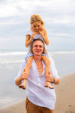 Babasının omuzlarında oturan bir kız. Baba ve kızı sahilde birlikte oynuyorlar. Mutlu bir aile. Birlikte eğleniyoruz. Kaliteli zaman. Yaz tatili. Asya 'da tatil. Aile bağları. Bali