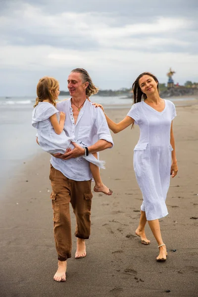Happy family concept. Summer holidays. Family vacation in Asia. Mother, father and daughter walking barefoot along the beach. Father carrying daughter in his arms. Copy space. Bali, Indonesia