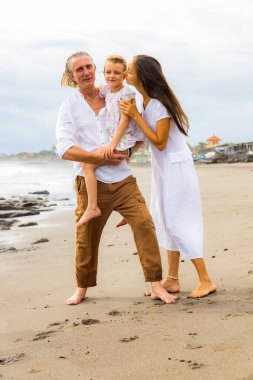 Kumsalda mutlu bir aile. Anne kızını öpüyor. Baba, kızını kollarında taşıyor. Birlikte güzel vakit geçirmek. Çıplak ayakla yürüyorum. Yaz tatili. Asya 'da tatil. Bali Adası, Endonezya