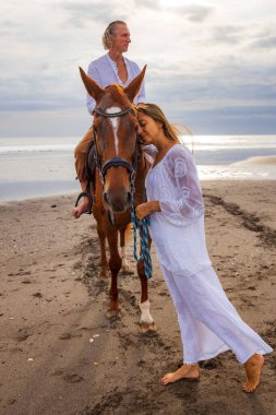 Sahilde ata binmek. Kahverengi atlı yakışıklı adam. Yanında duran ve bir atı okşayan bir kadın. Hayvanları sev. Karı koca birlikte vakit geçiriyorlar. Seçili odak. Bali, Endonezya