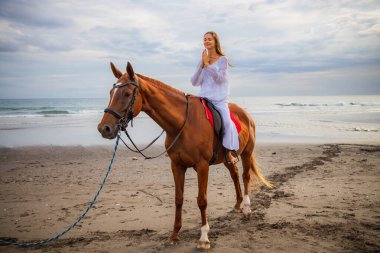 Kumsalda ata binen güzel beyaz kadın. Kapalı gözler. Eller namaste mudra 'da. Beyaz elbiseli bir kadın. Uzayı kopyala Sahilde gün batımı zamanı. Açık hava aktiviteleri. Bali, Endonezya