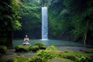 Yoga nilüfer pozu. Genç beyaz kadın taşın üzerinde oturuyor, meditasyon yapıyor, yoga yapıyor, şelalede pranayama yapıyor. Eller namaste mudra 'da. Yoga inzivası. Tibumana Şelalesi, Bali, Endonezya