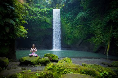 Yoga nilüfer pozu. Genç beyaz kadın taşın üzerinde oturuyor, meditasyon yapıyor, yoga yapıyor, şelalede pranayama yapıyor. Eller namaste mudra 'da. Yoga inzivası. Tibumana Şelalesi, Bali, Endonezya
