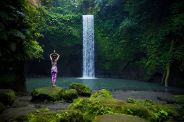 Kafkasyalı kadın taşın üzerinde duruyor, yoga yapıyor. Şelale yakınlarında namaste Mudra ile kol kaldıran genç bir kadın. Yoga yaşam tarzı. Tibumana şelalesi, Bali, Endonezya. Arkadan görüntüle.