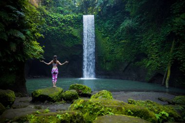 Kafkasyalı kadın taşın üzerinde duruyor, yoga yapıyor. Genç bir kadın şelalenin yanında Gyan Mudra ile kollarını kaldırıyor. Yoga yaşam tarzı. Tibumana şelalesi, Bali, Endonezya. Arkadan görüntüle.