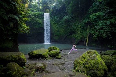 Şelalenin yanındaki yoga. Virabhadrasana I, Savaşçı I Poz veriyor. Asana ayakta duruyor. Yandan bak. Sağlıklı bir yaşam tarzı. Yoga inzivası. Tibumana şelalesi, Bali