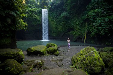 Açık havada yoga pratiği. Şelalenin yanında duran genç bir kadın. Eller namaste mudra 'da, gözler kapalı. Güneş selamı için hazırlanıyor Surya Namaskar. Sakin sakin. Yoga inzivası. Tibumana şelalesi, Bali