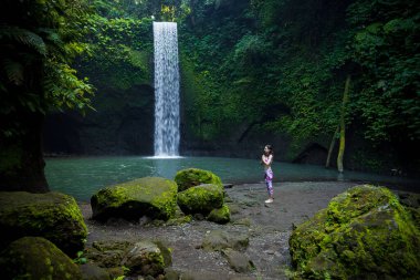 Açık havada yoga pratiği. Şelalenin yanında duran genç bir kadın. Eller namaste mudra 'da, gözler kapalı. Güneş selamı için hazırlanıyor Surya Namaskar. Sakin sakin. Yoga inzivası. Tibumana şelalesi, Bali