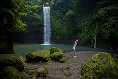 Yoga yapan sağlıklı genç kadın Surya Namaskar, Hasta Uttanasana 'ya şelale yakınlarında poz veriyor. Sağlıklı yaşam konsepti. Yoga inzivası. Tropik bir manzara. Tibumana şelalesi, Bali