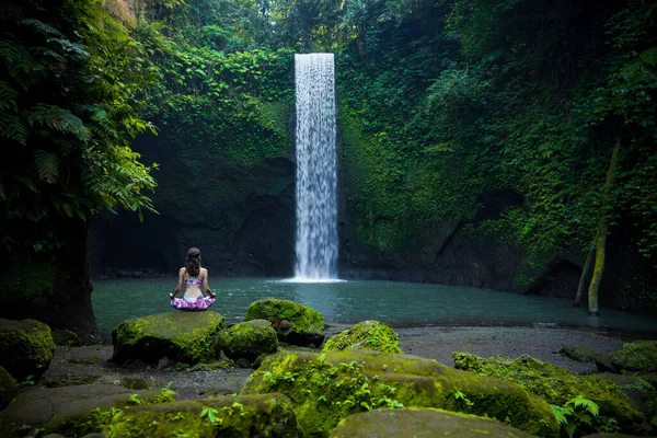 Yoga nilüfer pozu. Genç beyaz kadın taşın üzerinde oturuyor, meditasyon yapıyor, yoga yapıyor, şelalede pranayama yapıyor. Eller Gyan Mudra 'da. Yoga inzivası. Arkadan bak. Tibumana Şelalesi, Bali, Endonezya