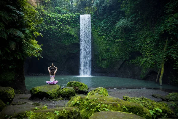 Yoga nilüfer pozu. Genç beyaz kadın taşın üzerinde oturuyor, yoga yapıyor, şelalede pranayama yapıyor. Namaste Mudra 'da eller havaya. Yoga inzivası. Arkadan bak. Tibumana şelalesi Bali