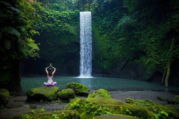 Yoga nilüfer pozu. Genç beyaz kadın taşın üzerinde oturuyor, yoga yapıyor, şelalede pranayama yapıyor. Namaste Mudra 'da eller havaya. Yoga inzivası. Arkadan bak. Tibumana şelalesi Bali