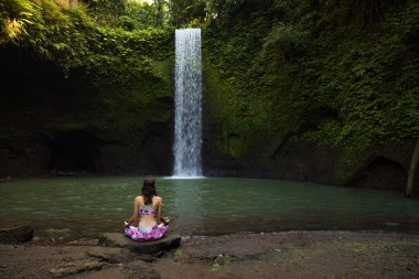 Yoga nilüfer pozu. Genç beyaz kadın taşın üzerinde oturuyor, meditasyon yapıyor, yoga yapıyor, şelalede pranayama yapıyor. Eller Gyan Mudra 'da. Yoga inzivası. Arkadan bak. Tibumana Şelalesi, Bali, Endonezya