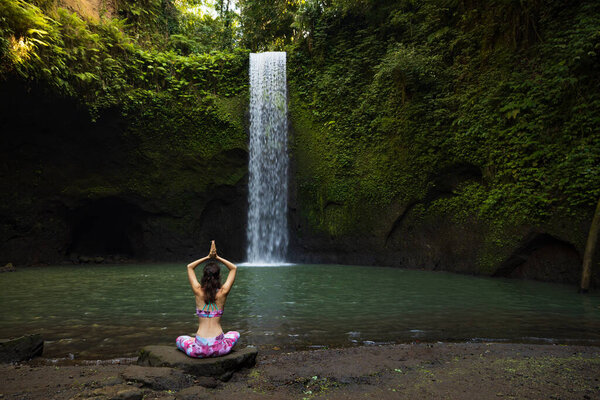 Yoga lotus pose. Young Caucasian woman sitting on the stone, meditating practicing yoga, pranayama at waterfall. Hands raised up in namaste mudra. Yoga retreat. View from back. Tibumana waterfall Bali