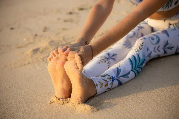 Close up of female feet. Woman sitting on the sand, practicing yoga. Paschimottanasana, Seated Forward Bend Pose. Hands holding big toes. Outdoor yoga concept. Working out. Thomas beach, Bali