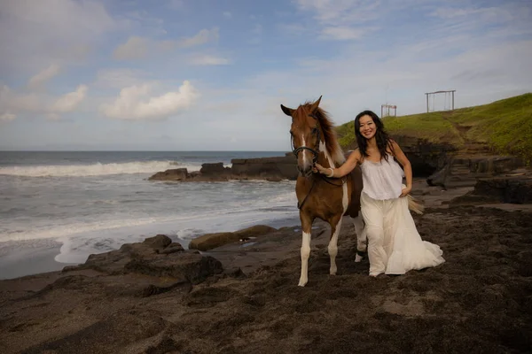 Happy woman leading horse by its reins. Horse riding on the beach. Human and animals relationship. Asian woman wearing long white dress. Nature concept. Copy space. Bali, Indonesia