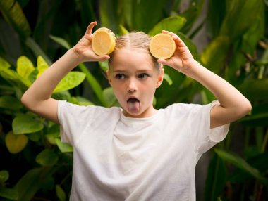 İki dilim taze limonu olan küçük bir kız. Beyaz kız limon tutuyor, komik surat yapıyor, dil gösteriyor. Seçili odak. Yeşil tropik yapraklar arka planda. Organik meyve konsepti. Bali, Endonezya