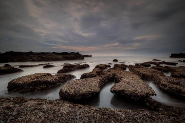 Arka plan için manzaralı deniz manzarası. Taşları ve taşları olan bir sahil. Dalgalar çekildi. Bulanık sisli su. Bulutlu gökyüzü. Kapama hızı yavaş. Yumuşak odaklanma. Uzayı kopyala Mengening Plajı, Bali, Endonezya
