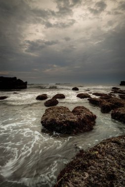 Arka plan için güzel bir deniz manzarası. Taşları ve taşları olan bir sahil. Dalgalar çekildi. Hareket suyu. Bulutlu gökyüzü. Kapama hızı yavaş. Yumuşak odaklanma. Uzayı kopyala Dikey yerleşim. Mengening Plajı, Bali, Endonezya