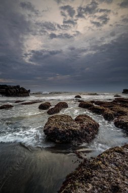 Arka plan için güzel bir deniz manzarası. Taşları ve taşları olan bir sahil. Dalgalar çekildi. Hareket suyu. Bulutlu gökyüzü. Kapama hızı yavaş. Yumuşak odaklanma. Uzayı kopyala Dikey yerleşim. Mengening Plajı, Bali, Endonezya