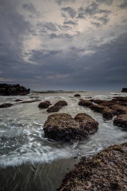 Arka plan için güzel bir deniz manzarası. Taşları ve taşları olan bir sahil. Dalgalar çekildi. Hareket suyu. Bulutlu gökyüzü. Kapama hızı yavaş. Yumuşak odaklanma. Uzayı kopyala Dikey yerleşim. Mengening Plajı, Bali, Endonezya