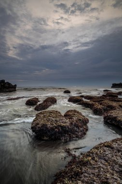 Arka plan için güzel bir deniz manzarası. Taşları ve taşları olan bir sahil. Dalgalar çekildi. Hareket suyu. Bulutlu gökyüzü. Kapama hızı yavaş. Yumuşak odaklanma. Uzayı kopyala Dikey yerleşim. Mengening Plajı, Bali, Endonezya