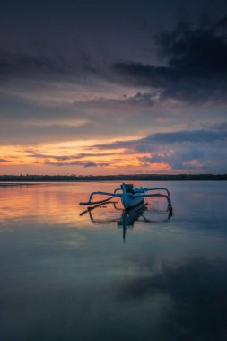 Deniz Burnu. Balıkçı teknesi. Geleneksel Balili tekne jukung. Gün doğarken sahilde balıkçı teknesi. Su yansıması. Bulutlu gökyüzü. Ufukta güneş ışığı görünüyor. Kapama hızı yavaş. Yumuşak odaklanma. Sanur, Bali