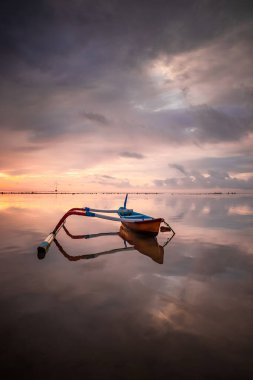 Deniz Burnu. Balıkçı teknesi. Geleneksel Balili tekne jukung. Gün doğarken sahilde balıkçı teknesi. Su yansıması. Bulutlu gökyüzü. Ufukta güneş ışığı görünüyor. Kapama hızı yavaş. Yumuşak odaklanma. Sanur, Bali