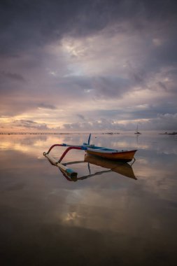 Deniz Burnu. Balıkçı teknesi. Geleneksel Balili tekne jukung. Gün doğarken sahilde balıkçı teknesi. Su yansıması. Bulutlu gökyüzü. Ufukta güneş ışığı görünüyor. Kapama hızı yavaş. Yumuşak odaklanma. Sanur, Bali