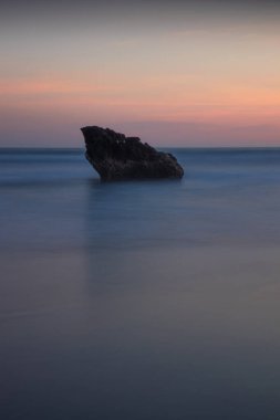 Gün batımı deniz manzarası. Okyanusta taş gibi. Kapama hızı yavaş. Yumuşak odaklanma. İpeksi su. Renkli gökyüzü. İnanılmaz bir su yansıması. Doğa ve çevre geçmişi. Uzayı kopyala Endonezya, Bali Plajı.