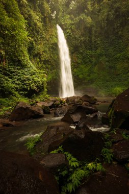 Şelale manzarası. Tropikal yağmur ormanlarında saklı güzel bir şelale. Büyük taşlı ön plan. Yavaş deklanşör hızı, hareket fotoğrafçılığı. Seyahat ve macera. Nung Nung Şelalesi, Bali, Endonezya