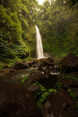Şelale manzarası. Tropikal yağmur ormanlarında saklı güzel bir şelale. Büyük taşlı ön plan. Yavaş deklanşör hızı, hareket fotoğrafçılığı. Seyahat ve macera. Nung Nung Şelalesi, Bali, Endonezya