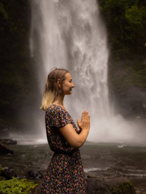 Genç beyaz kadın şelale yakınlarında vakit geçiriyor. Meditasyon ve rahatlama. Eller namaste mudra 'da. Kapalı gözler. Seyahat yaşam tarzı. Elbise giyen kadın. Endonezya, Bali 'de Nung Nung Şelalesi
