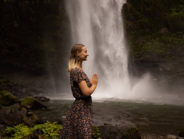 Genç beyaz kadın şelale yakınlarında vakit geçiriyor. Meditasyon ve rahatlama. Eller namaste mudra 'da. Kapalı gözler. Seyahat yaşam tarzı. Elbise giyen kadın. Endonezya, Bali 'de Nung Nung Şelalesi